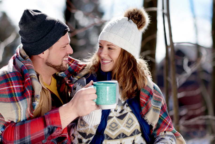 A man and a woman sitting outside in toques and sweaters, smiling at each other while the man holds up a warm drink